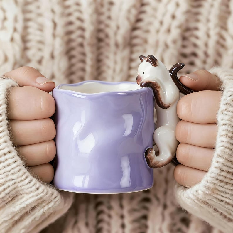 Puede incluir: Una taza de cer&aacute;mica lavanda con un asa en forma de gato. El gato es blanco con marcas marrones. La taza tiene un borde ondulado. La taza est&aacute; sostenida por manos que llevan un su&eacute;ter color crema.
