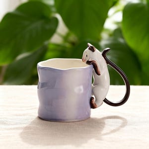 Puede incluir: Una taza de cer&aacute;mica con un degradado de color lila claro y blanco. Una figura de gato blanco y marr&oacute;n est&aacute; unida al costado, como si estuviera trepando. El asa es una cola marr&oacute;n curvada. La taza est&aacute; sobre una superficie neutra.