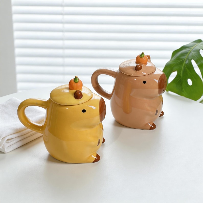 May include: Two ceramic mugs shaped like pigs, one yellow and one tan, each with a lid topped with a small pumpkin. The mugs have a handle and are sitting on a white surface. The mugs are designed with a cartoonish style.