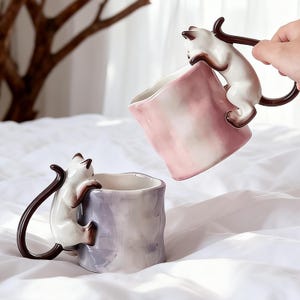 Puede incluir: Dos tazas de cer&aacute;mica con dise&ntilde;os de gatos. Las tazas son blancas, marrones y rosas, con un gato trepando por el lateral para formar el asa. Las tazas est&aacute;n sobre una superficie blanca.