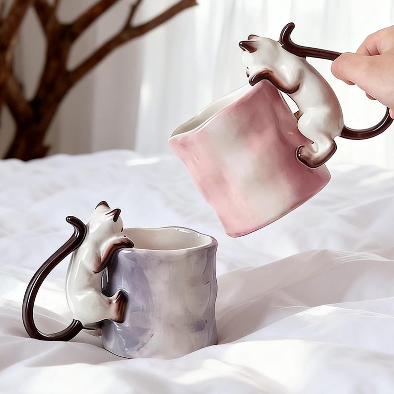 Puede incluir: Dos tazas de cer&aacute;mica con dise&ntilde;os de gatos. Las tazas son blancas, marrones y rosas, con un gato trepando por el lateral para formar el asa. Las tazas est&aacute;n sobre una superficie blanca.