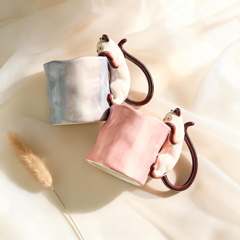 Puede incluir: Dos tazas de cer&aacute;mica, una azul claro y otra rosa, cada una con un asa en forma de gato blanco. Los gatos parecen estar trepando por las tazas. Las tazas tienen una forma texturizada y org&aacute;nica, sobre un fondo blanco suave.