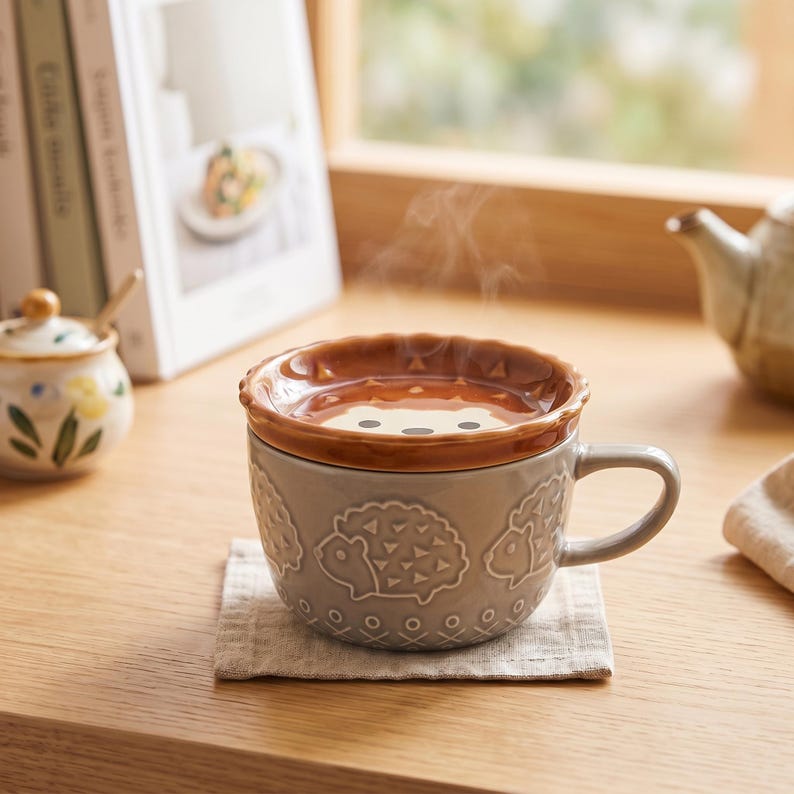 May include: A light grey ceramic mug featuring embossed hedgehog patterns around the exterior, accompanied by a matching brown lid with a stylised hedgehog face design. Steam rises from the mug, which rests on a square beige fabric coaster.