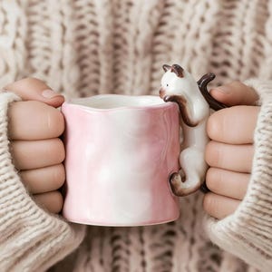 Puede incluir: Una taza de cer&aacute;mica rosa y blanca con un asa en forma de gato. La taza la sostiene una persona que lleva un su&eacute;ter color crema. El asa del gato es blanca con detalles marrones.
