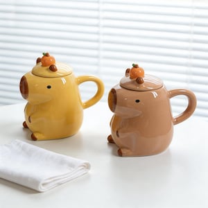 May include: Two ceramic mugs shaped like capybaras, one yellow and one brown, each with a lid topped with a small orange fruit. The mugs have a handle and small feet. A white folded cloth is in the foreground.