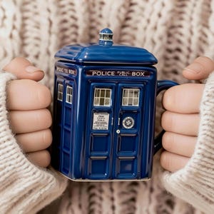 May include: A blue ceramic mug in the shape of a police call box, complete with a lid. The mug is inscribed with "POLICE PUBLIC BOX CALL" and has window and door details. The mug is held by someone wearing a cream-colored jumper.