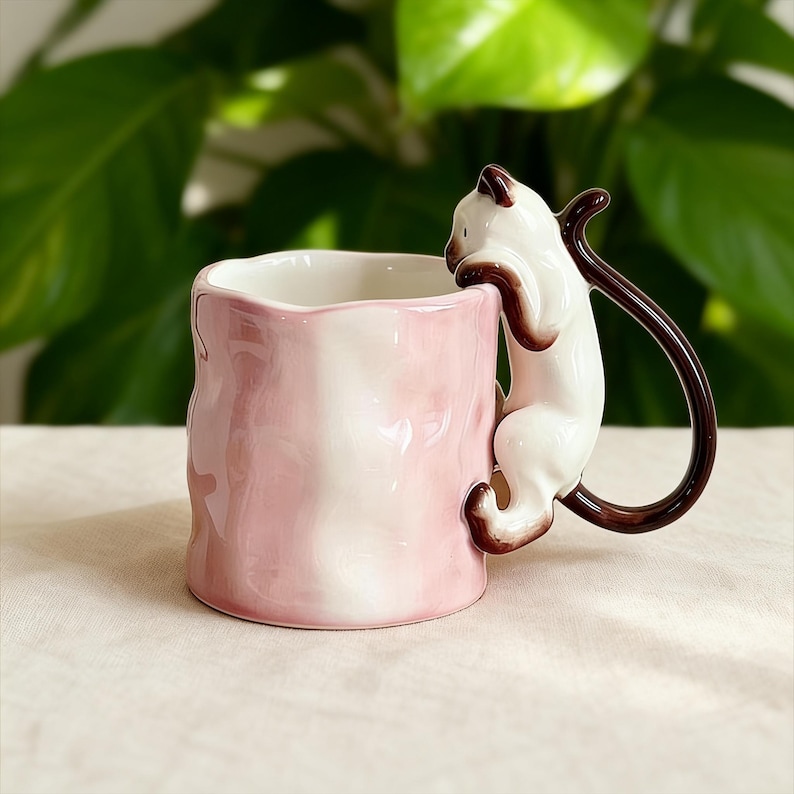 Puede incluir: Una taza de cer&aacute;mica rosa y blanca con un dise&ntilde;o &uacute;nico. La taza presenta un asa de gato siam&eacute;s esculpido, con el cuerpo y la cola del gato formando el asa. La taza tiene un exterior texturizado y un interior blanco.
