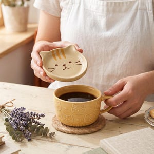 May include: A yellow ceramic mug with a matching lid featuring a cat face design. The mug body is embossed with a repeating pattern of cat faces, paw prints, and fish. The lid is shaped like a cat head with brown stripes and whiskers.