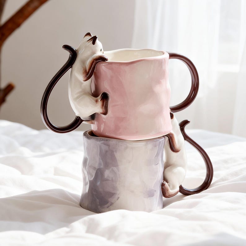 Puede incluir: Dos tazas de cer&aacute;mica apiladas. La taza superior es rosa y la inferior es lavanda. Cada taza tiene un asa marr&oacute;n y un gato blanco trepando por el lateral. Los gatos tienen orejas y colas marrones.