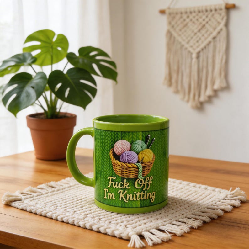Może przedstawiać: Zielony ceramiczny kubek z uchwytem, ​​przedstawiający kosz z kulkami przędzy i napisem "Fuck Off I'm Knitting". Kubek stoi na kremowej podkładce na drewnianym stole. Roślina doniczkowa i makrama ścienna są w tle.