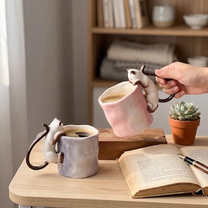 Puede incluir: Dos tazas de cer&aacute;mica con dise&ntilde;o de gato. Una taza es lavanda, la otra rosa. Cada taza tiene un gato trepando por el lateral, con la cola formando el asa. Ambas tazas est&aacute;n llenas de caf&eacute;. Tambi&eacute;n se ve una suculenta en una maceta de terracota y un libro abierto.