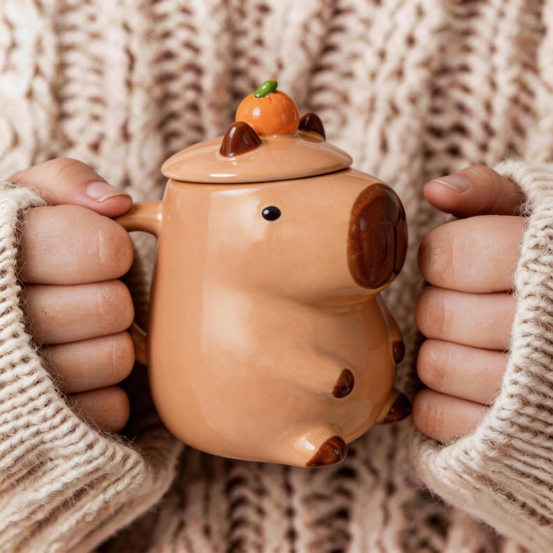 May include: A tan ceramic mug shaped like a capybara, with a matching lid topped with a small orange fruit. The mug has brown accents and a black eye. The mug is held by a person wearing a cream-colored sweater.