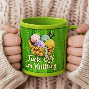 Może przedstawiać: Zielony ceramiczny kubek z uchwytem, przedstawiający kosz z kulkami przędzy i napisem "Fuck Off I'm Knitting". Kubek ma tło z wzorem dzianiny i jest trzymany przez osobę w kremowym swetrze.