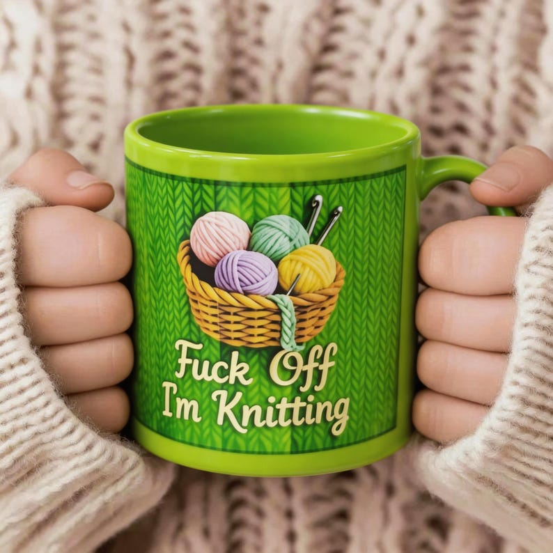 Może przedstawiać: Zielony ceramiczny kubek z uchwytem, przedstawiający kosz z kulkami przędzy i napisem "Fuck Off I'm Knitting". Kubek ma tło z wzorem dzianiny i jest trzymany przez osobę w kremowym swetrze.