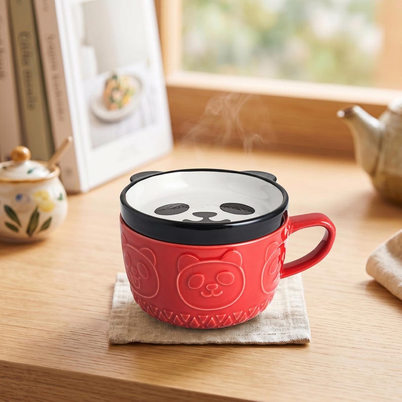 May include: A bright red ceramic mug featuring embossed panda bear faces around the exterior. The mug includes a matching lid with a black rim and a panda face design on top. Steam rises from the mug, which has a sturdy handle on the right side.