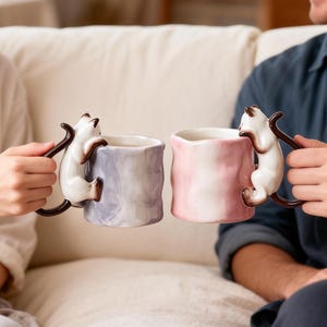 Puede incluir: Dos tazas de cer&aacute;mica, una lavanda y otra rosa, cada una con un gato blanco trepando por el lateral como asa. Las tazas son sostenidas por dos personas. Las asas de los gatos son marrones.