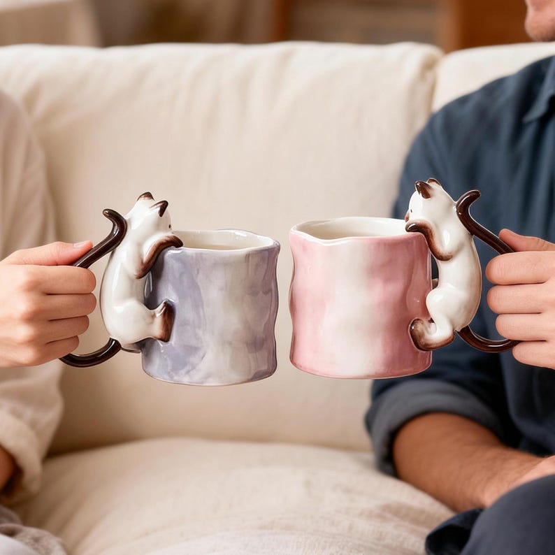Puede incluir: Dos tazas de cer&aacute;mica, una lavanda y otra rosa, cada una con un gato blanco trepando por el lateral como asa. Las tazas son sostenidas por dos personas. Las asas de los gatos son marrones.