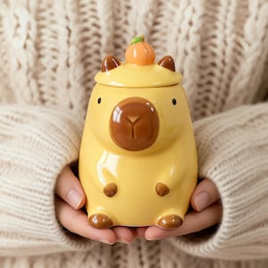 May include: A yellow ceramic mug shaped like a capybara, with brown accents and a pumpkin lid. The mug has a glossy finish and is held in a person's hands. The mug is approximately 4 inches tall.