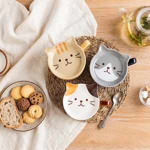 May include: Three ceramic mugs shaped like cat heads, featuring a tan tabby design, a grey and white bicolor design, and a white, orange, and brown calico design. Each mug has a handle, cat ears on the rim, and a printed face with whiskers.