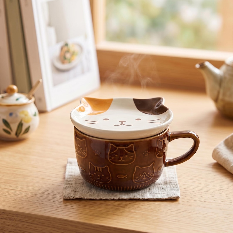 May include: A brown ceramic mug with a handle and a matching lid featuring a calico cat face design. The mug body is decorated with embossed cat faces and small fish icons. Steam rises from the lid, which has white, orange, and brown patches.
