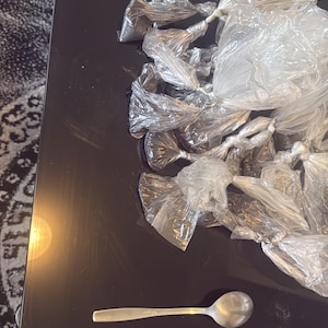 Puede incluir: Primer plano de una mesa de vidrio negro con un montón de pequeñas bolsas de plástico blancas atadas con nudos. Una cuchara de plata está sobre la mesa.