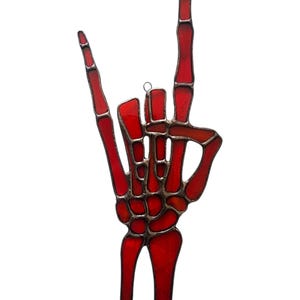 May include: A stained glass decoration in the shape of a hand making the 'rock on' gesture. The hand is a vibrant red color with a detailed skeletal design. The glass is outlined with a dark, metallic border.