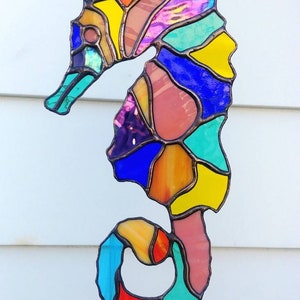 May include: A colorful stained glass seahorse with a blue, yellow, orange, pink, and green color scheme. The seahorse is hanging from a chain.