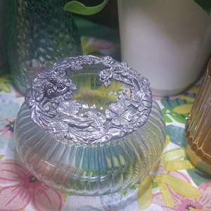 May include: A clear glass jar with a silver floral design lid. The jar has a ribbed design and is perfect for storing small items.