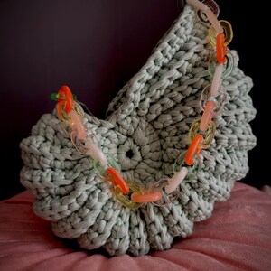 Peut inclure: Un sac crocheté vert clair avec une chaîne colorée. Les maillons de la chaîne sont transparents et présentent des teintes orange, jaune et rose. Le sac repose sur une surface rose, avec un fond sombre.