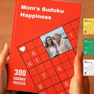 Peut inclure: Un livre rouge avec le titre "Mom's Sudoku Happiness" et une photo de deux personnes sur la couverture. Le livre contient 300 puzzles Sudoku.