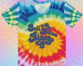 May include: A tie-dye t-shirt with a v-neck design. The shirt features a vibrant spiral pattern in shades of blue, yellow, and red. The text "Bell Bottom Blues" is displayed in a denim-style font. The shirt is a casual wear item.