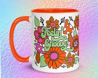 May include: White ceramic mug with an orange handle and interior. The mug features a colorful floral design with the words "Feelin' Groovy!" in green and orange. The design includes flowers, mushrooms, and leaves.