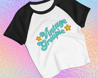 May include: White and black raglan t-shirt with the text "Vintage Groupie" in turquoise script, accented with orange flowers. The shirt has a crew neck and short sleeves.