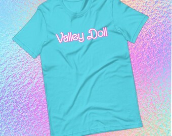May include: A turquoise t-shirt with the text "Valley Doll" in pink script. The short-sleeved shirt is made of a soft material and is displayed against a colorful, iridescent background.