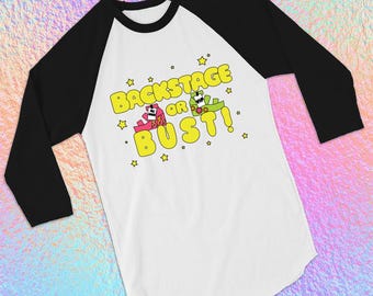 May include: White and black raglan baseball tee with the text "BACKSTAGE OR BUST!" in yellow. The graphic includes two cartoon characters in pink and green, surrounded by yellow stars.