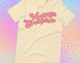 May include: Cream-colored t-shirt with the text "Vintage Groupie" in pink, outlined in a darker pink. The design features a heart symbol. The shirt has short sleeves and a crew neck.