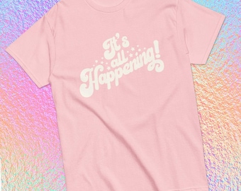 May include: A light pink t-shirt with the phrase "It's all Happening!" in a white, retro-style font. The text is surrounded by small white stars. The shirt is made of a soft material and has short sleeves.
