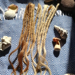 Peut inclure: Deux ensembles de dreadlocks synthétiques, l'un blond et l'autre brun, disposés sur une surface à motifs bleus et blancs.