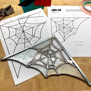 May include: A stained glass spider web pattern with 29 pieces, measuring 10.5 inches by 10 inches. The pattern is printed on white paper and includes numbered pieces for assembly. The finished stained glass spider web is also shown in the image, measuring 7.5 inches by 7.25 inches and consisting of 21 pieces.