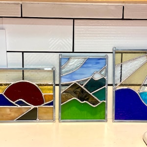 May include: Three stained glass panels depicting different landscapes. The first panel shows a red sun setting behind blue mountains with a gold sky. The second panel shows green mountains with a blue sky and white clouds. The third panel shows a blue sky with a yellow sun and blue water.
