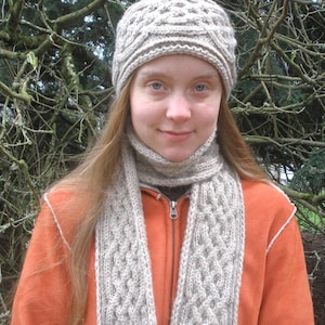 May include: A woman wearing a light brown knitted hat and matching scarf. The hat and scarf have a cable knit pattern. She is wearing a bright orange jacket.