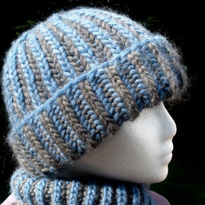 Può includere: Un cappello di lana blu e grigio con una sciarpa abbinata. Il cappello è morbido e ha una trama a coste.