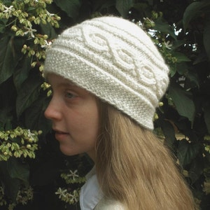 May include: A white knitted hat with a cable knit pattern. The hat is worn by a person with long, light brown hair.