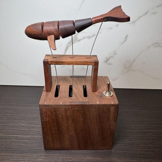 A hand-carved walnut whale that actually swims in a wave motion when cranked — kinetic automata meets desk sculpture meets music box.