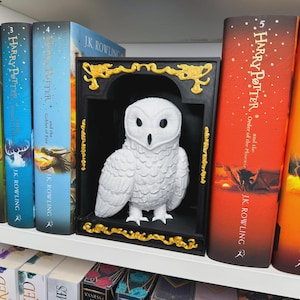Könnte beinhalten: Eine weiße Eulenfigur in einem schwarzen und goldenen dekorativen Rahmen, platziert auf einem weißen Regal neben mehreren "Harry Potter"-Büchern. Die Eule hat detaillierte Federstrukturen. Der Rahmen hat goldene Akzente.