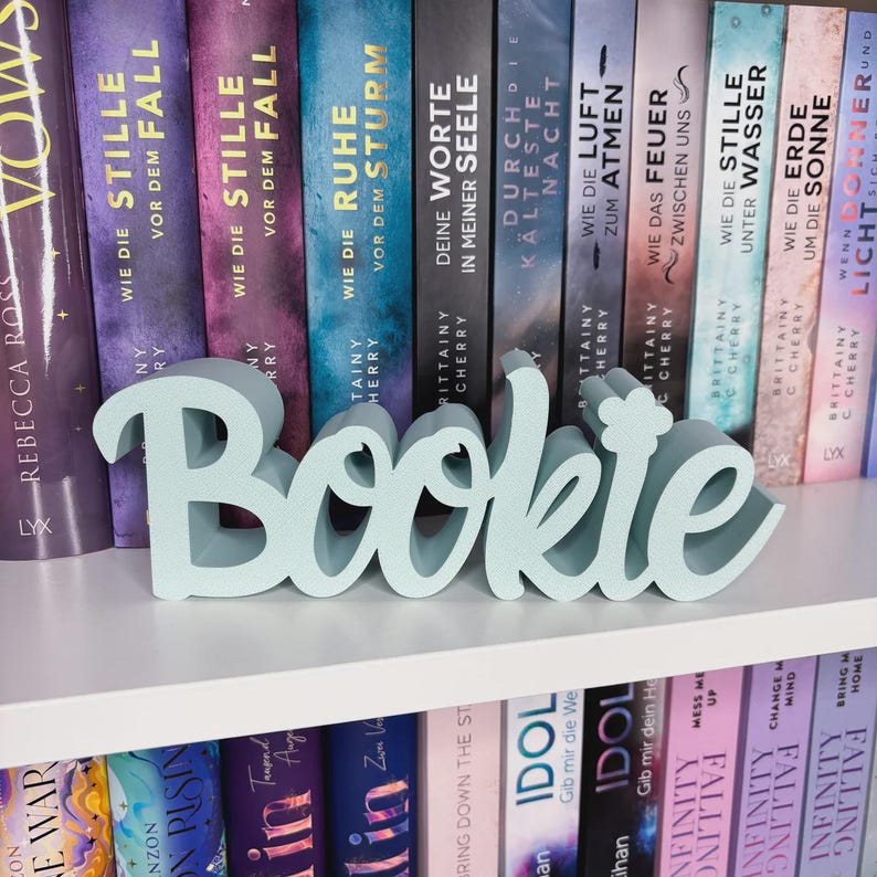 Op de afbeelding: Een lichtblauw houten "Bookie" bord in een sierlijk lettertype staat op een witte plank voor een rij boeken met Duitse titels. Het bord is een decoratief item voor boekenliefhebbers.