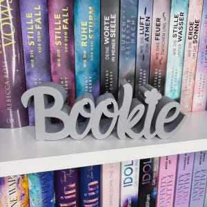 Op de afbeelding: Een grijs 'Bookie' woordkunst decoratie-object in cursief schrift staat op een witte plank voor een rij kleurrijke boeken. De boeken hebben Duitse titels. Een decoratief item voor boekenliefhebbers.