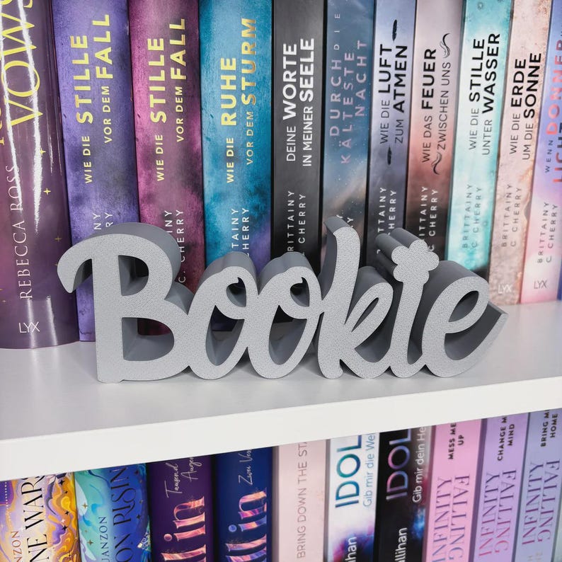 Op de afbeelding: Een grijs 'Bookie' woordkunst decoratie-object in cursief schrift staat op een witte plank voor een rij kleurrijke boeken. De boeken hebben Duitse titels. Een decoratief item voor boekenliefhebbers.