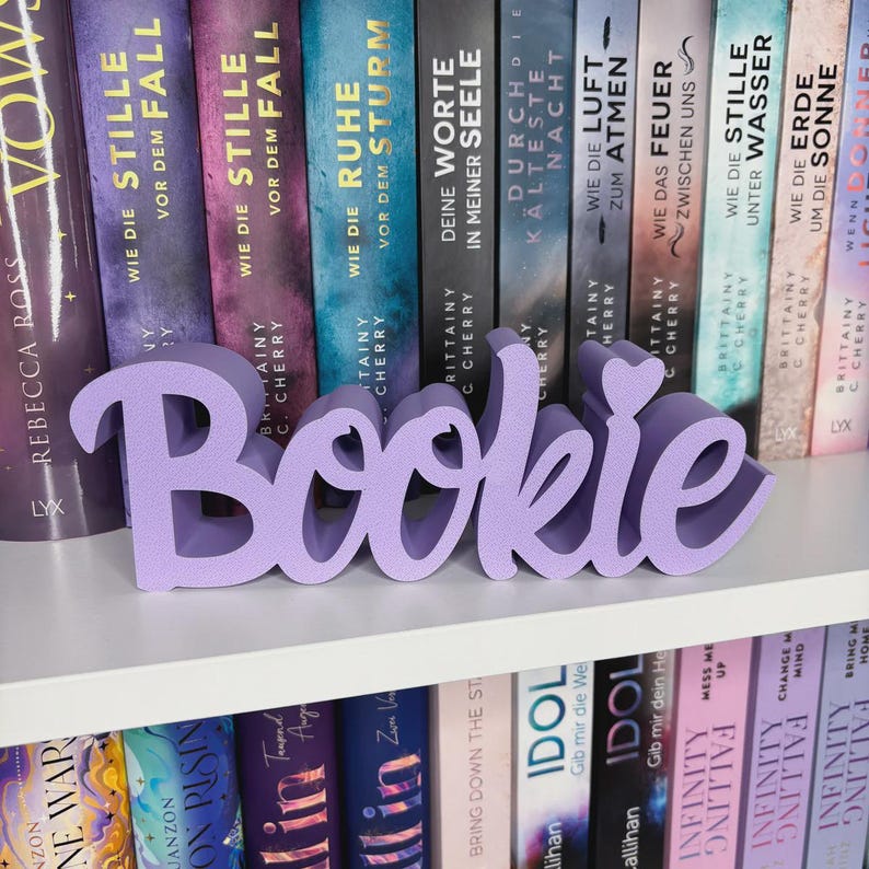 Op de afbeelding: Een lavendelkleurig "Bookie" woordkunstwerk staat op een witte plank voor een rij boeken. Het woord is in een cursief lettertype. De boeken hebben kleurrijke ruggen met titels in verschillende talen.