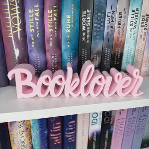 Op de afbeelding: Een roze "Booklover" woordkunst decoratie staat op een witte plank voor een rij boeken. De boeken hebben kleurrijke ruggen met titels en auteursnamen. De woordkunst is in een cursief lettertype.